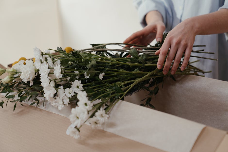 A person arranging a bouquet of fresh white flowers, including chrysanthemums and sweet peas, with green foliage such as eucalyptus, on a light-colored wooden surface. The bouquet is partially wrapped