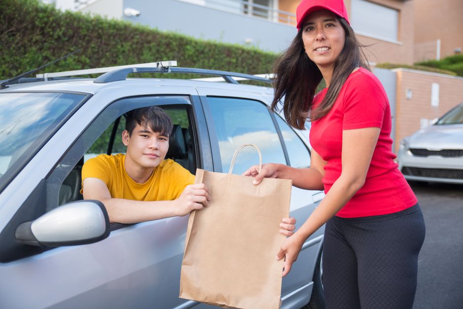 A woman in a red shirt and matching cap hands over a brown paper shopping bag to a young man sitting in the driver's seat of a silver car. The young man, wearing a yellow t-shirt, reaches out to accep