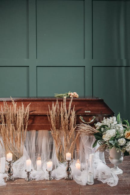 A floral arrangement featuring a bouquet of white roses, peonies, and greenery placed on top of a polished wooden casket against a green paneled wall. In front of the casket, there are tall, beige dri