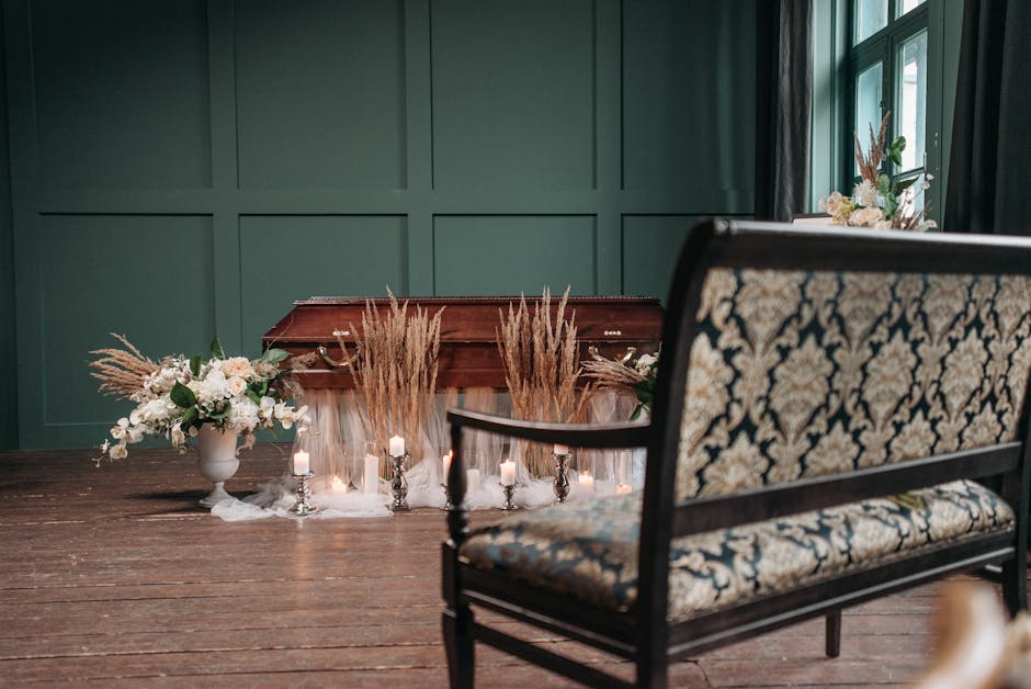 A floral arrangement displayed on a table against a dark wooden paneled wall features a bouquet of white roses, lilies, and greenery in a white vase, accompanied by tall dried pampas grass. Several sm