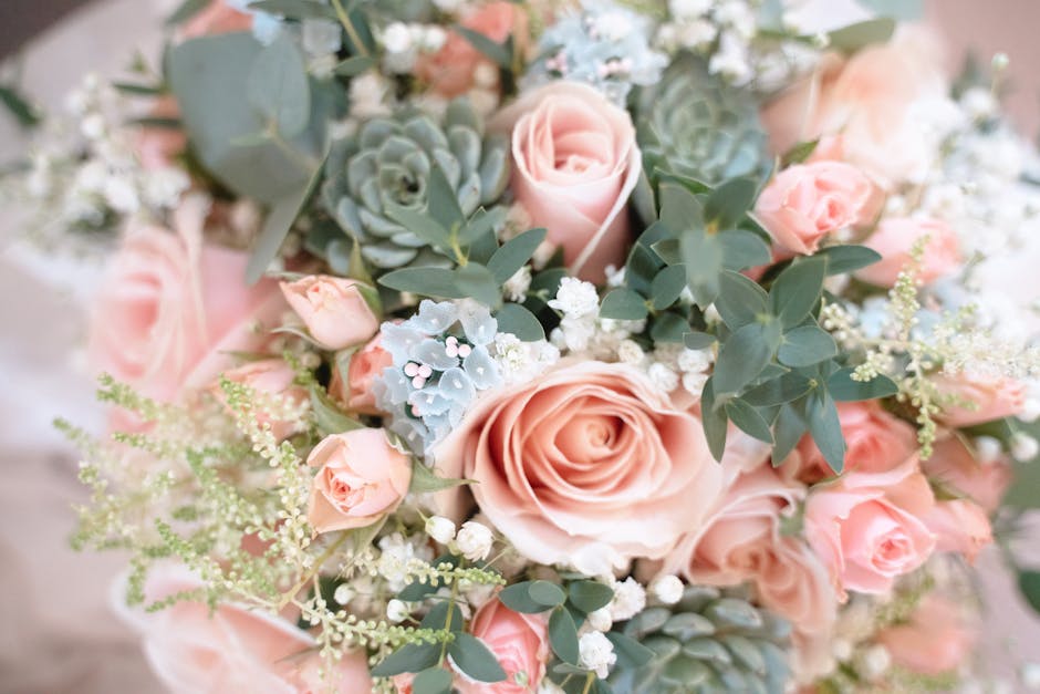 A floral arrangement featuring a bouquet of soft pink roses, complemented by clusters of small white filler flowers and dusty green eucalyptus leaves. The bouquet is arranged in a rounded, lush style 