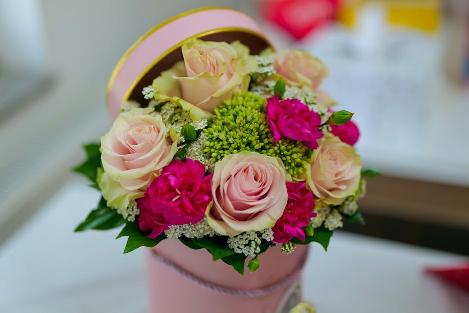 A round floral gift box featuring a carefully arranged bouquet of fresh flowers, including soft pink roses, vibrant pink carnations, clusters of small white blooms, and green foliage. The flowers are 