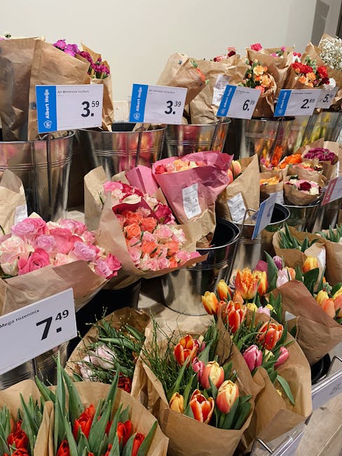 A display of fresh floral bouquets arranged in cylindrical metal buckets and wrapped in brown paper at a flower shop. The bouquet in the foreground features vibrant red and peach tulips with green fol
