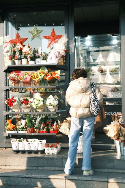 A woman stands outside a flower shop, looking at a display of floral arrangements and bouquets. The shop features clear glass shelves filled with colorful flowers, including red, white, and orange blo